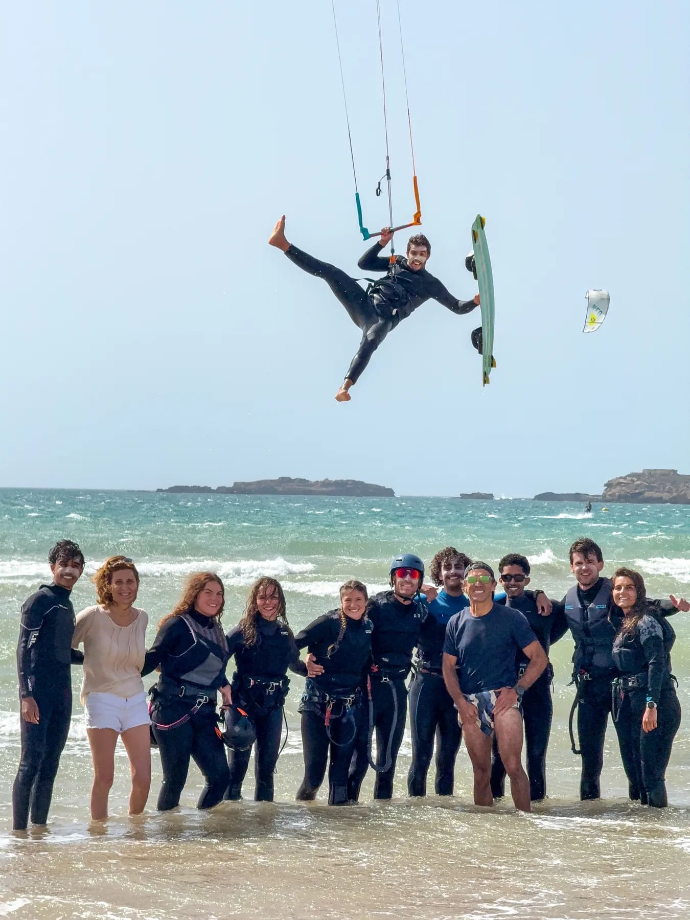 Stage de kitesurf à Essaouira — groupe d'élèves dans l'eau et rider en plein saut au-dessus d'eux, planche à la main, îles de Mogador à l'horizon, Windy Essaouira