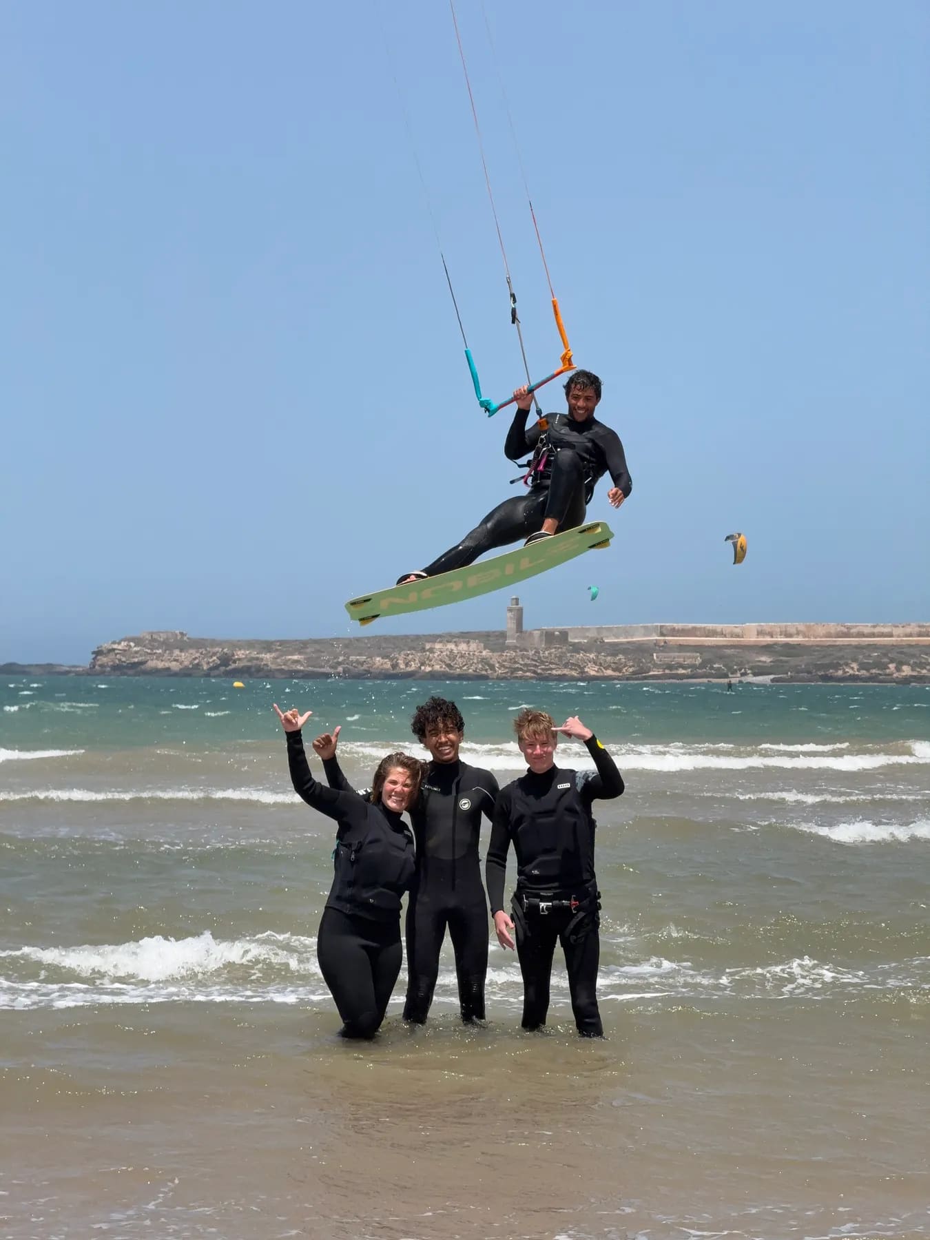 Kitesurf à Essaouira — rider en plein saut devant trois élèves qui l'encouragent depuis l'eau, île de Mogador en arrière-plan, Windy Essaouira