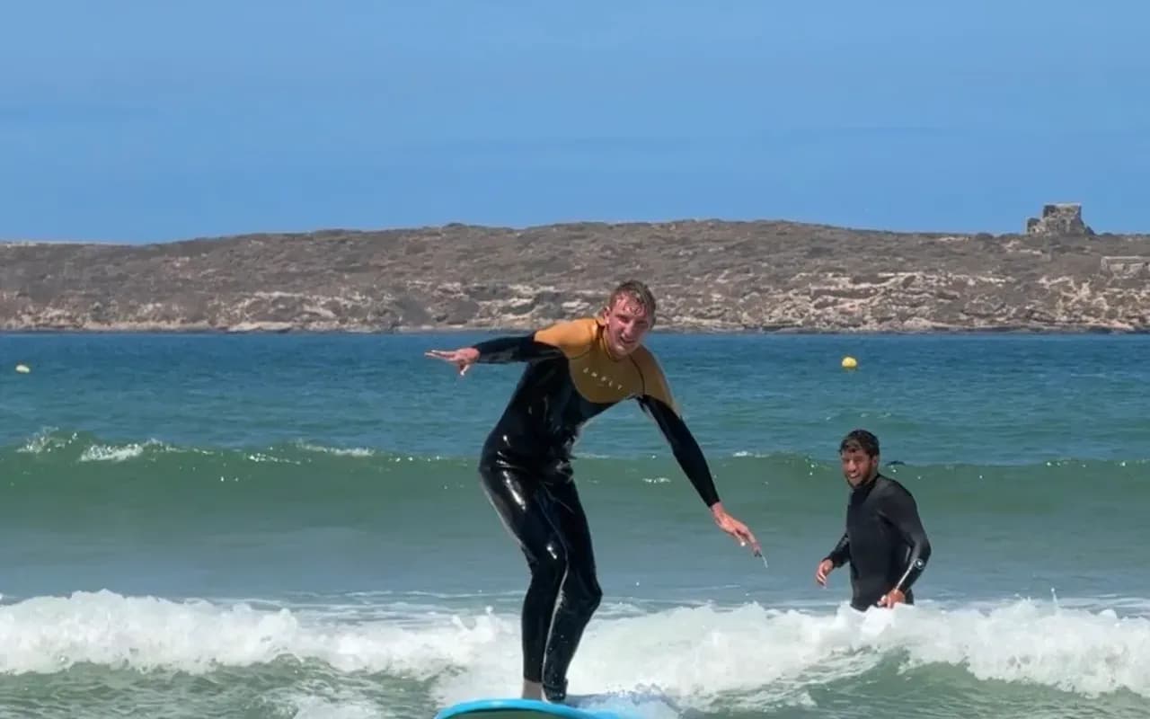 Élève debout sur sa planche dans la mousse à Essaouira, le moniteur juste à côté dans l'eau