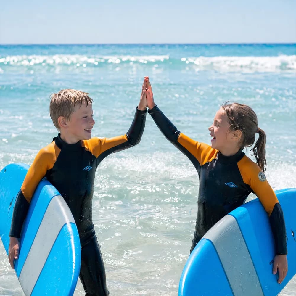 Deux enfants en combinaison se tapent dans la main, planches de surf en mousse sous le bras