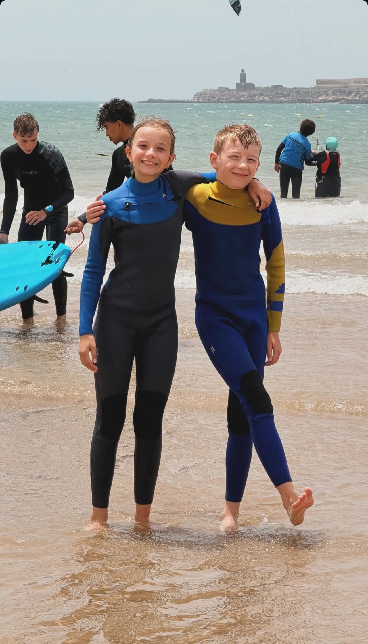 Deux enfants souriants en combinaison sur la plage d'Essaouira après leur cours de surf, autres élèves et moniteur derrière — Windy Essaouira